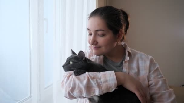 Młoda brunetka z kotem wygląda przez okno — Wideo stockowe