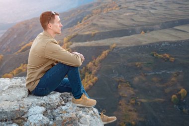 Genç adam sonbahar ağaçlarının arka planında bir kayanın üzerinde oturuyor.