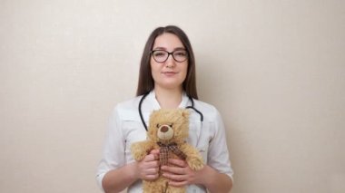 Gözlüklü kadın çocuk doktoru, oyuncak ayıyı bej renkte tutuyor.