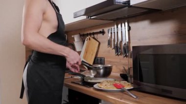 Çıplak gövdeli, siyah önlüklü, tanınmayan bir adam mutfakta krep hazırlıyor.