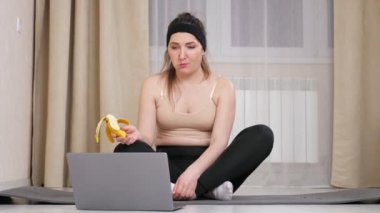 Spor kıyafetli bir kadın laptopa bakar ve muz yer.