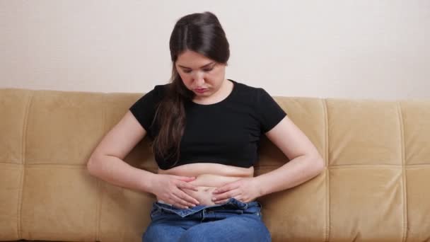 Fiatal nő érzés kövér redők a gyomorban, miközben ül a kanapén — Stock videók