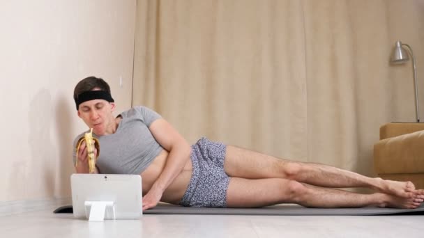 Pria feminin makan pisang, melihat tablet dan melambaikan kaki dengan canggung saat berbaring di lantai — Stok Video