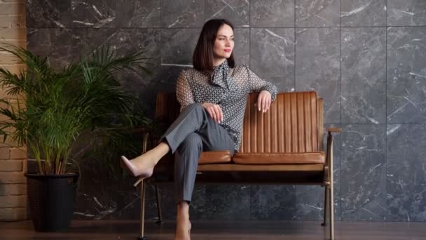 Mladá brunetka v šedém klasickém kostýmu sedí na hnědé lavičce — Stock video