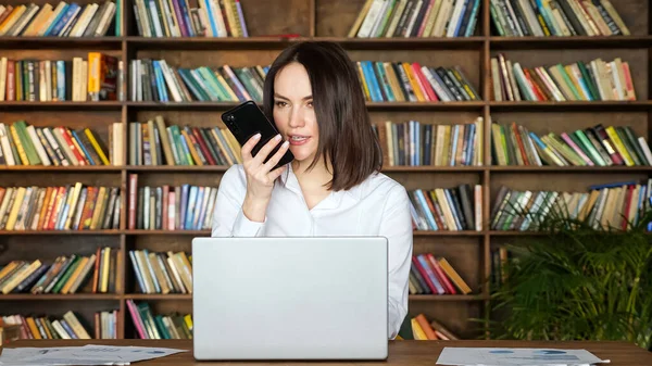 Brunette woman talks on phone near laptop at table in room