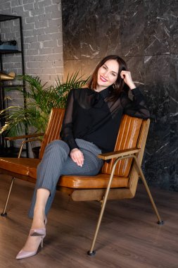 Esmer kadın, resmi kıyafetlerle deri koltukta oturuyor. Bir ofis dolabının bir çeşidi.