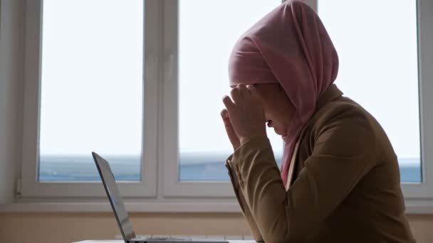 Donna in hijab rosa prende una pausa e massaggia il ponte del naso mentre si lavora su un computer portatile — Video Stock