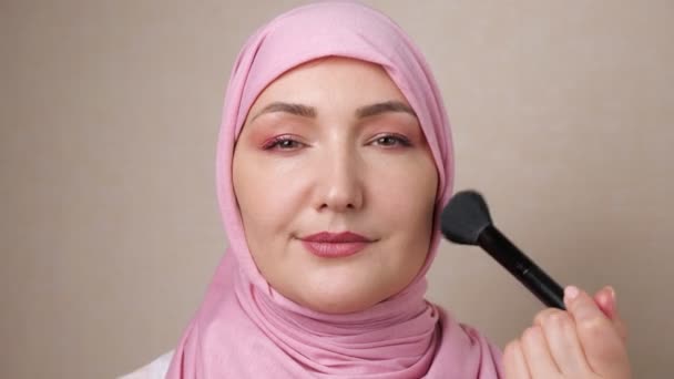 Woman in hijab strokes face with a soft brush while smiling at the camera — Stock Video