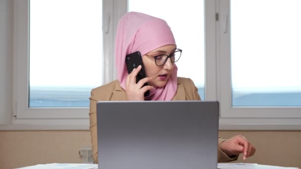 Женщина в хиджабе изучает документы и работает на ноутбуке, разговаривая по телефону — стоковое видео
