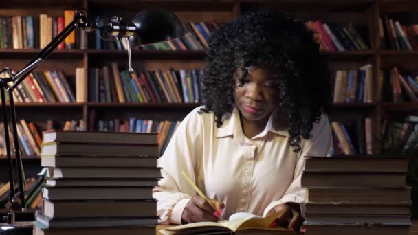 Senhora afro-americana escreve perto de pilhas de livros à mesa — Vídeo de Stock