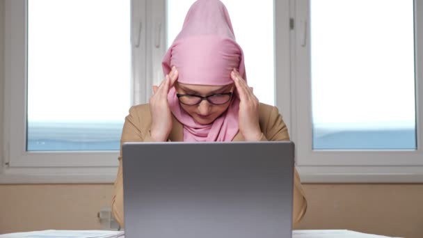 Γυναίκα σε μια μαντίλα κρατά το κεφάλι της και μασάζ ναούς, ενώ εργάζονται σε ένα φορητό υπολογιστή — Αρχείο Βίντεο
