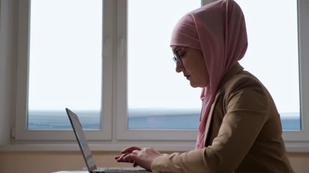 Mujer en hijab rosa escribiendo en el ordenador portátil contra el fondo de la ventana — Vídeo de stock