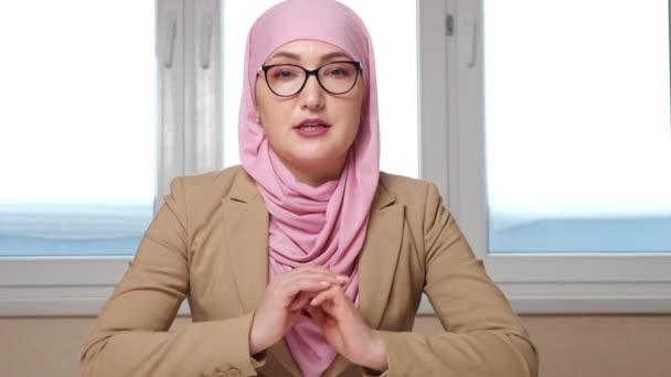 Mujer en hijab y gafas se sienta a la mesa y habla con la cámara, alejándose de la cámara — Vídeo de stock