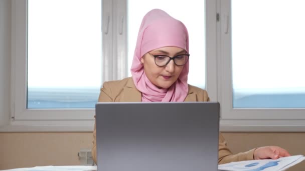 Mujer musulmana en pañuelo rosa para la cabeza y gafas de trabajo en el portátil y con documentos — Vídeo de stock