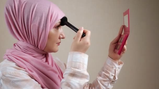 Joven mujer musulmana en hijab aplicando maquillaje con un cepillo mirando en el espejo — Vídeo de stock