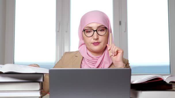 Giovane donna musulmana in hijab rosa che parla per videochiamata su laptop tra pile di libri — Video Stock