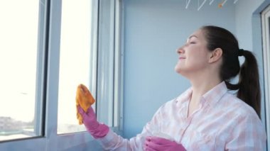 Eldivenli, sprey ve paçavralı genç bir kadın pencereyi temizliyor.