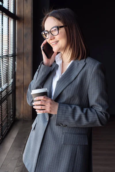 Деловая женщина с кофе стоит у окна и говорит по телефону — стоковое фото