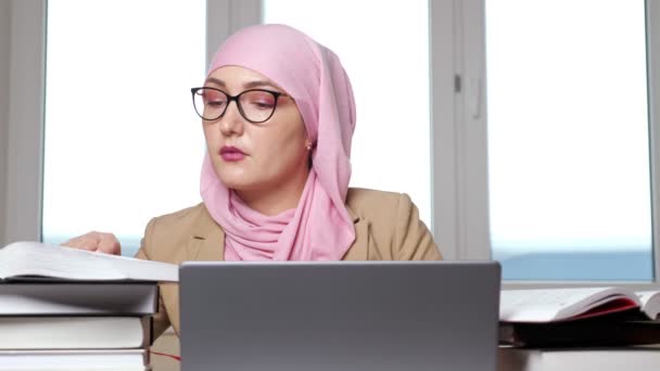 Mujer en hijab y gafas escribiendo en el portátil mirando en los libros — Vídeo de stock