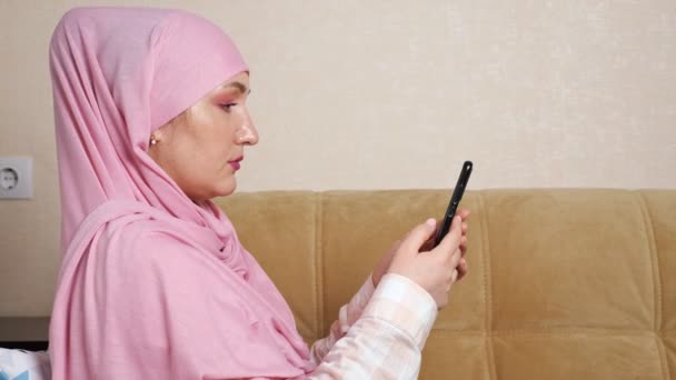 Wanita muslim muda dalam kerudung merah muda duduk di sofa dengan telepon — Stok Video