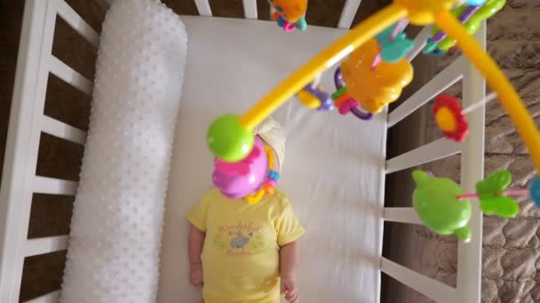 Alegre niña en amarillo se encuentra en cuna blanca con móvil — Vídeo de stock