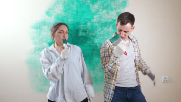 Joyful lady and guy sing into paintbrush as mics near wall — Stock video