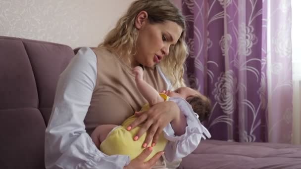 Η νεαρή μητέρα κουνάει το μωρό στην αγκαλιά της ενώ κάθεται στον καναπέ. — Αρχείο Βίντεο