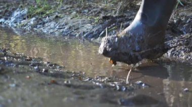 Lastik çizmeli tanınmaz bir adam çamurlu bir su birikintisine adım atıyor, ağır çekim.