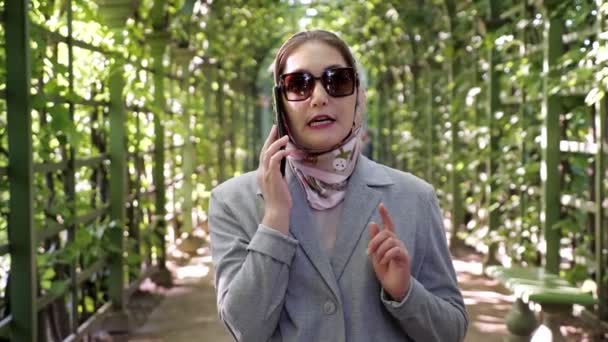 Σοβαρή επιχειρηματίας με μαντίλα και γυαλιά ηλίου μιλώντας στο τηλέφωνο στην αψίδα του κήπου — Αρχείο Βίντεο