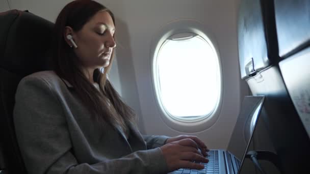 Une femme brune en costume et un écouteur sans fil dans l'oreille tapent du texte sur un ordinateur portable alors qu'elle est assise dans un avion — Video