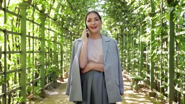 Wanita muda berbicara di telepon dalam koridor melengkung pohon di taman — Stok Video