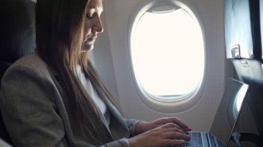 Takım elbiseli genç bir kadın uçağın camında dizüstü bilgisayarında yazı yazıyor.