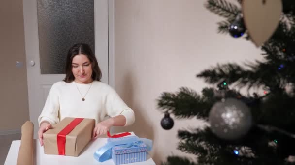 Femme brune emballe des cadeaux à la table à côté d'un sapin décoré — Video