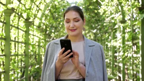 Junge Frau in grauer Jacke tippt Text am Telefon im Gartenbogen — Stockvideo