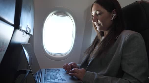 Morena em terno com fone de ouvido sem fio na orelha funciona no laptop enquanto sentado no avião — Vídeo de Stock