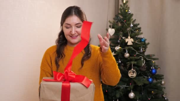 Woman in a mustard sweater unties a red ribbon from a gift box on the background of a decorated christmas tree — Stock Video