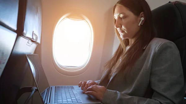Brunette woman in a suit and a wireless earphone in ear is typing text on a laptop while sitting on an airplane