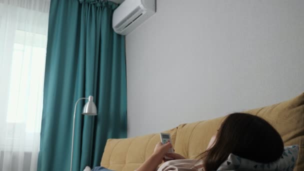 Αγνώριστη γυναίκα ανοίγει το κλιματιστικό με το τηλεχειριστήριο, ξαπλωμένη στον καναπέ — Αρχείο Βίντεο