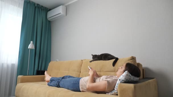 Woman turns on the air conditioner by the remote control, lying on the couch with a cat — Stock Video