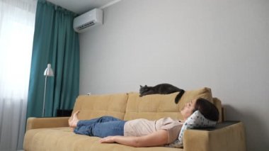 Genç bir kadın ve kedi klimalı bir odada kanepede yatıyorlar.