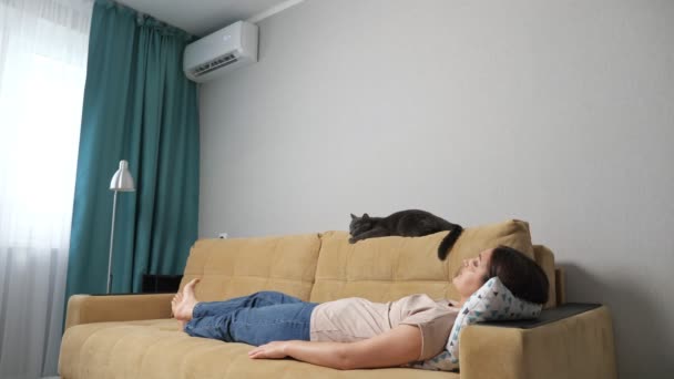 Junge Frau und Katze liegen auf einem Sofa in einem Raum mit Klimaanlage — Stockvideo