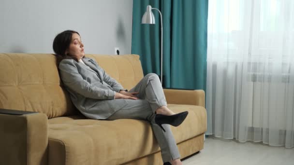 Mujer joven en un traje formal se sienta en el sofá quitándose los zapatos. Masaje de pies, hinchazón de pies — Vídeos de Stock