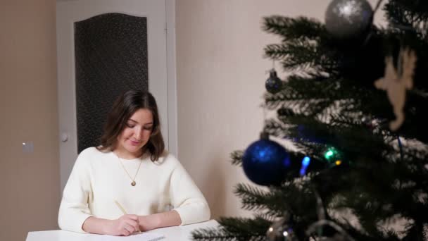 Ung kvinna skriver med penna på papper bredvid julgranen — Stockvideo