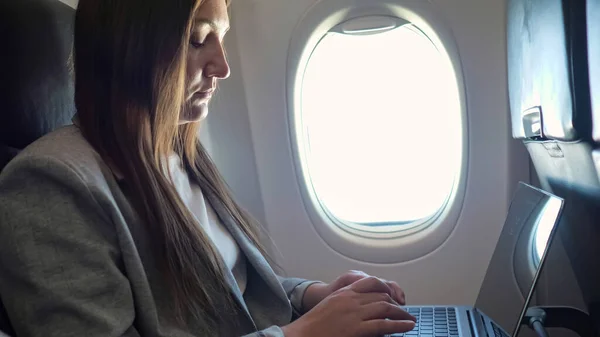 Молодая женщина в костюме печатает текст на ноутбуке у окна в самолете — стоковое фото