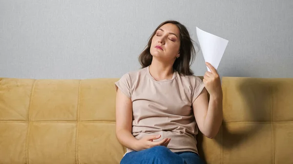 Junge Frau, die selbst unter Hitze leidet, sitzt mit einem Blatt Papier auf der Couch — Stockfoto