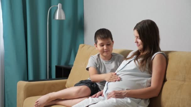 Těhotná matka a syn na gauči. Chlapec hladí a objímá břicho — Stock video