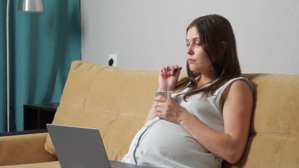 Young pregnant woman eating yogurt and looking at laptop while sitting on sofa — Stock Video
