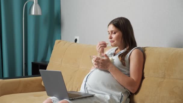 Zwangere vrouw op zoek naar laptop en het eten van popcorn terwijl zitten op de bank — Stockvideo