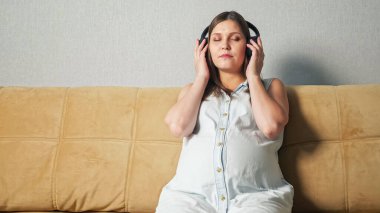 Kanepede otururken kulaklık takan telefonlu hamile bir kadın.