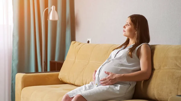 Jeune femme enceinte caressant le ventre tout en étant assis sur le canapé — Photo